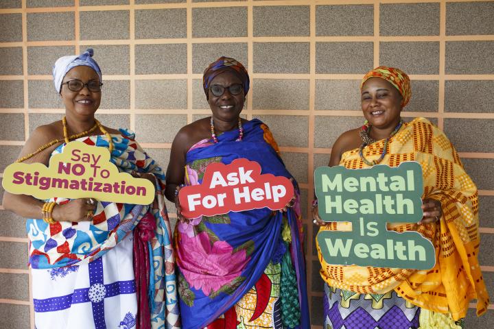  Drei Frauen schauen in die Kamera und lachen. Sie halten Schilder hoch, auf denen in Englisch steht: Sag Nein zu Stigmatisierung; Frage nach Hilfe; Mentale Gesundheit ist Reichtum.  